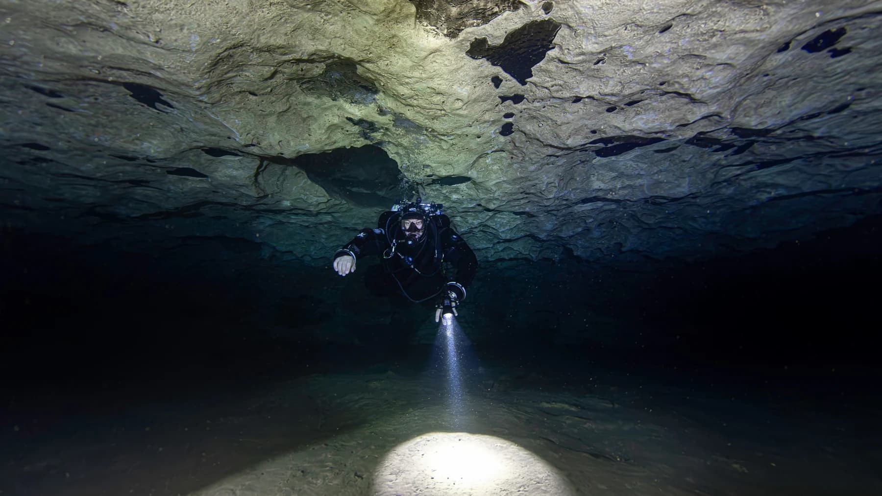A cave diver beneath a limestone ceiling, illuminating the passage with a primary light